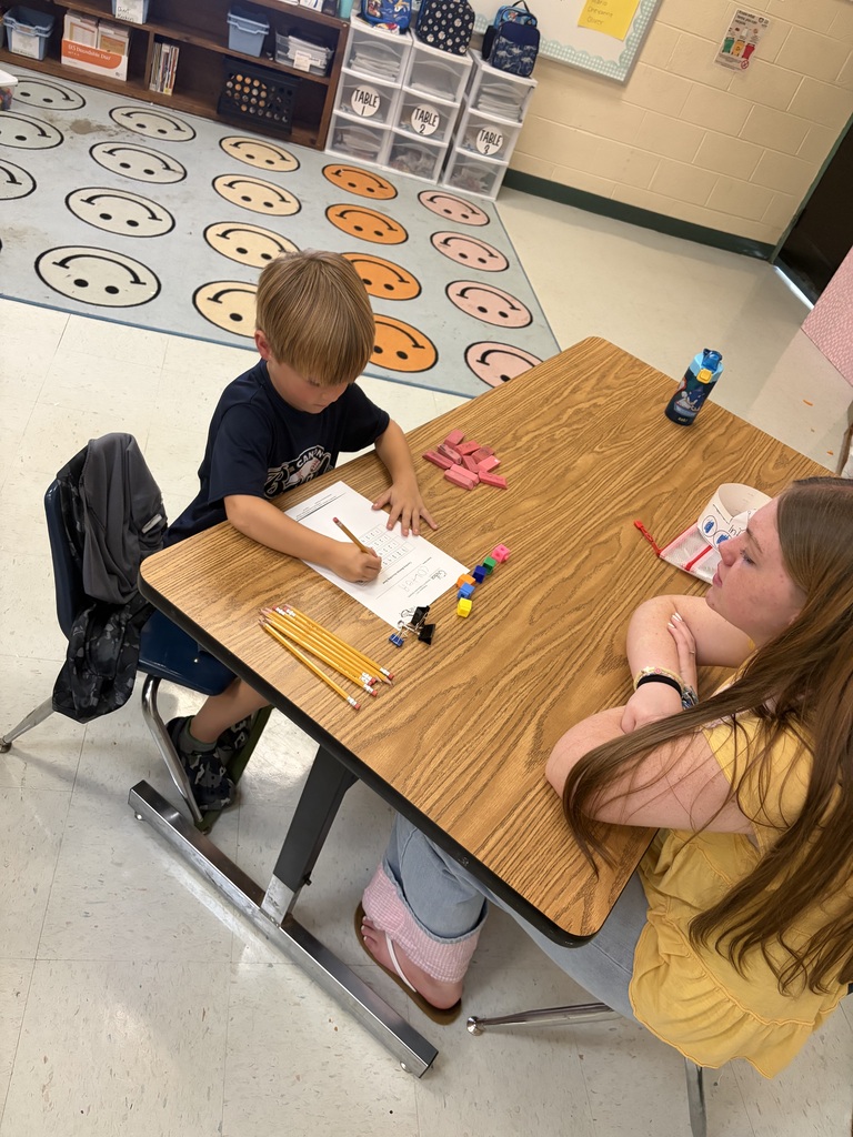 A child and an adult sit at a wooden table, working on a school task. Background includes a patterned floor and a storage unit.