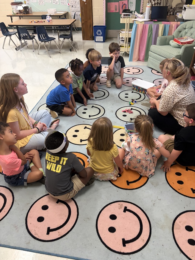 Group of young children sitting on a rug with smiley faces, facing an adult who appears to be reading to them.