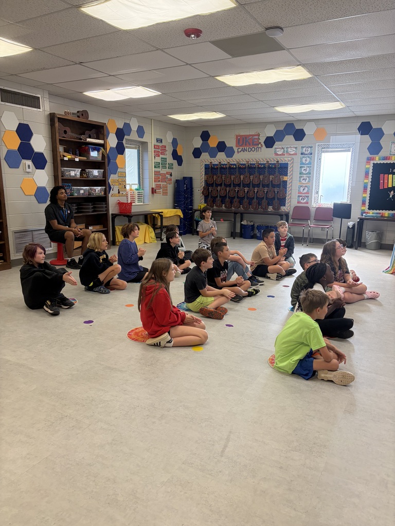 Group of children sitting on floor, some wearing sneakers. One woman stands at the front. Hexagon patterns on walls.