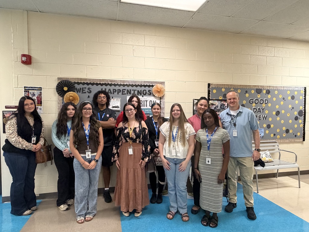 A group of people in a room with a blue rug, white walls, and bulletin boards.