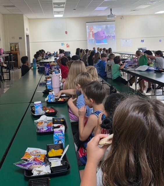 A classroom with children seated at tables. Trays with food, utensils, and snacks are on the tables.