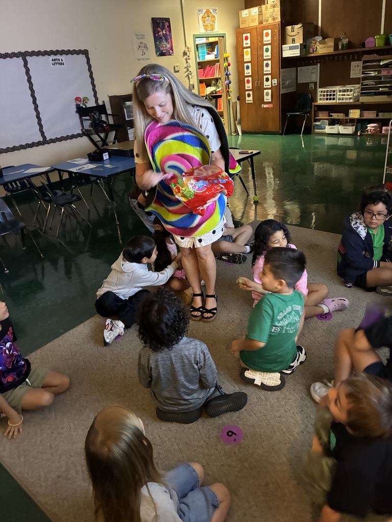 A woman with a large candy swirl stands with children sitting on a rug in a classroom.