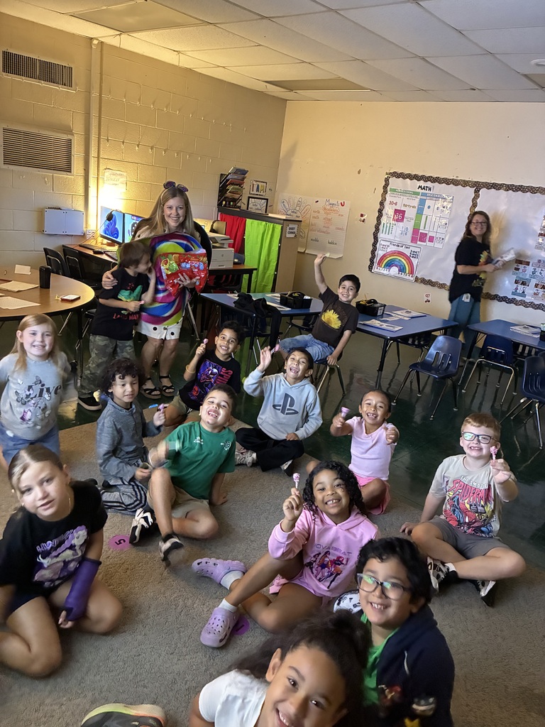 Group of children in a classroom with some sitting on the floor. Some are smiling and posing for a photo.