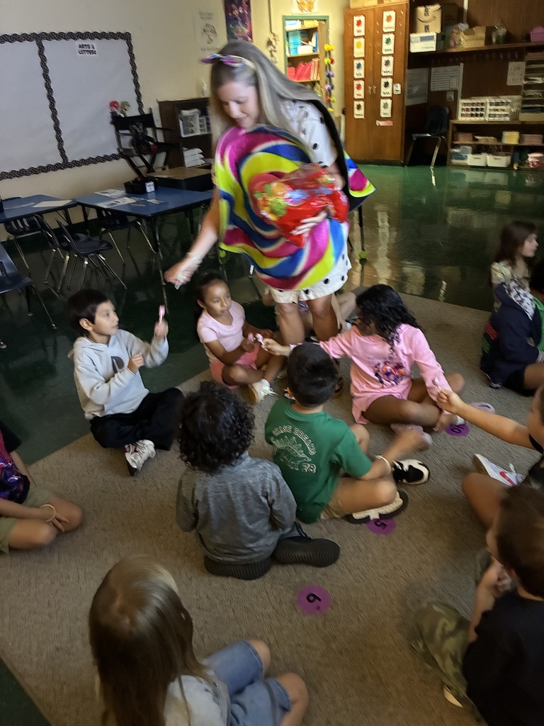 A woman in a colorful outfit stands before a group of children in a classroom.