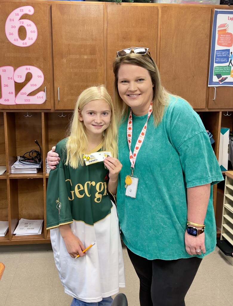 A woman and a girl, both smiling, stand in a room. The girl holds a certificate, and the woman wears a green shirt and a lanyard.