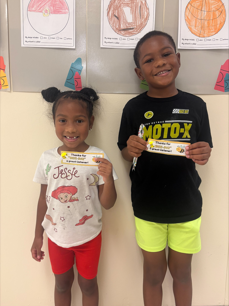 Two children standing next to each other, one with a yellow certificate, posing in front of a white wall with colorful drawings.