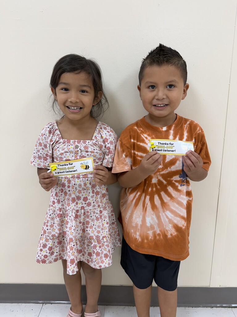 Two children stand close, one with a pink dress and the other in a tie-dye shirt, holding certificates.