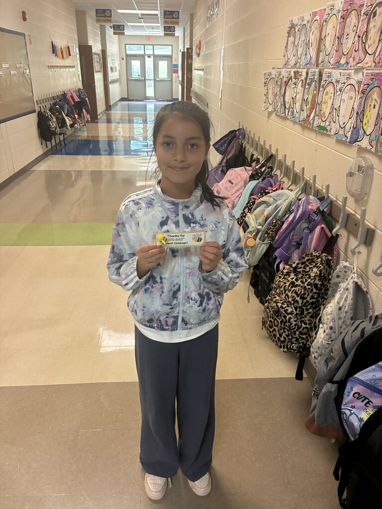 A girl with a yellow item stands in a school hallway with bags on hooks and art on walls.