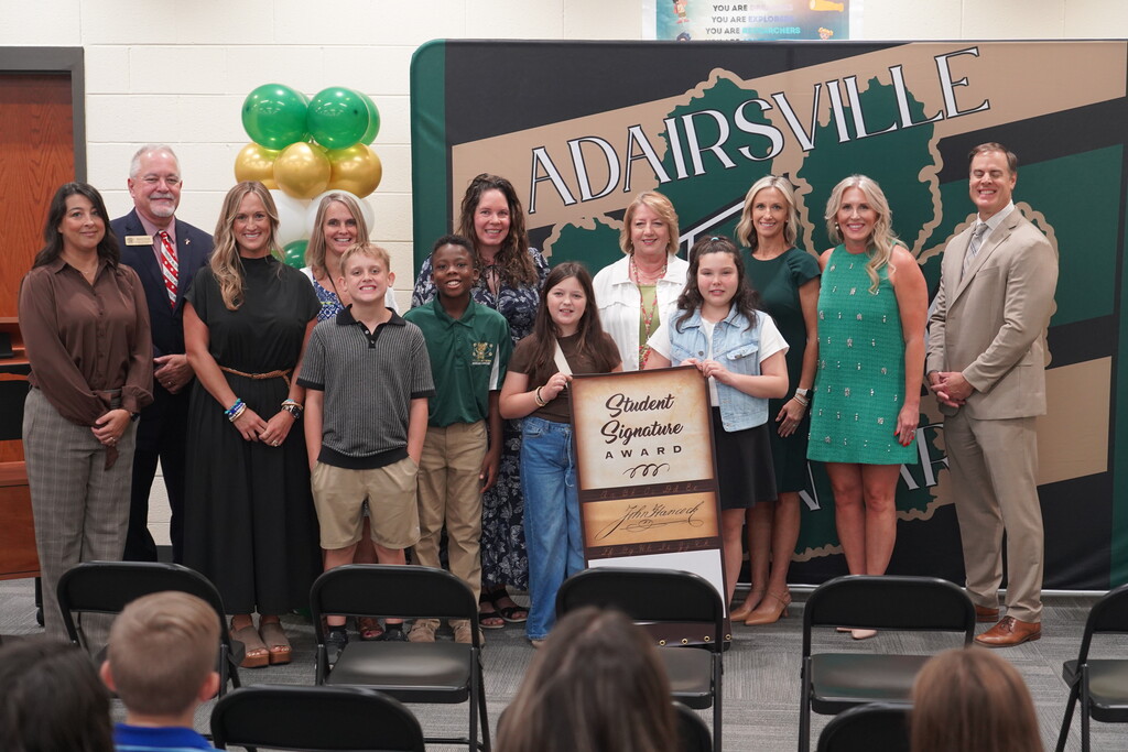 A group of adults and children pose with a certificate. Behind them is a banner reading "Adairsville".