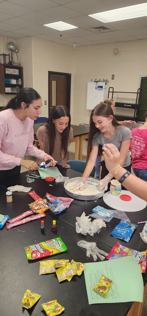 A group of people work together at a table with various food items, supplies, and papers.