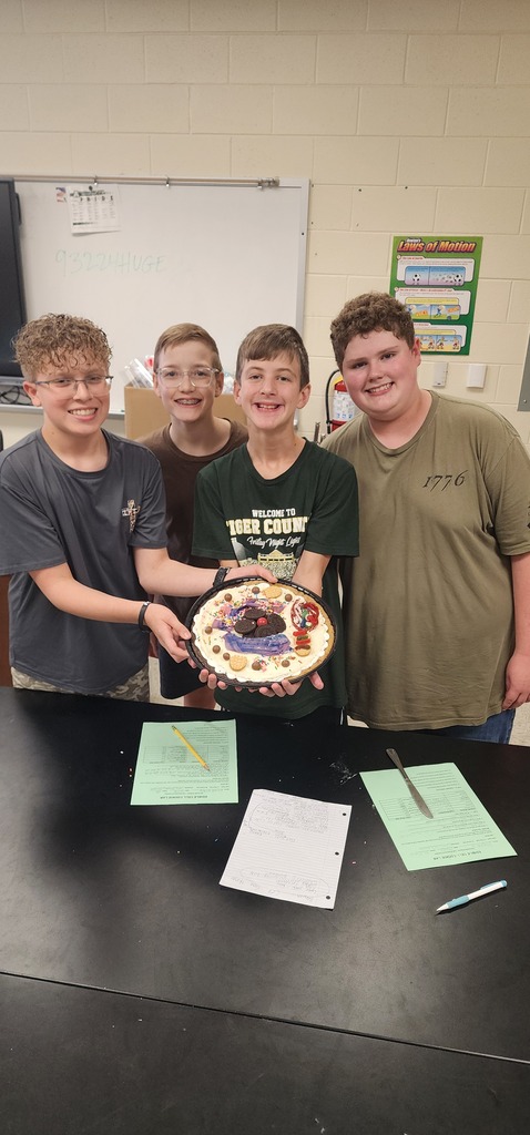 Four young boys stand at a table, holding a circular cake. Papers and a pen are on the table.