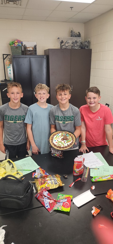 Four boys stand around a table with snacks, a trophy, and papers. A bag and some papers are on the table.