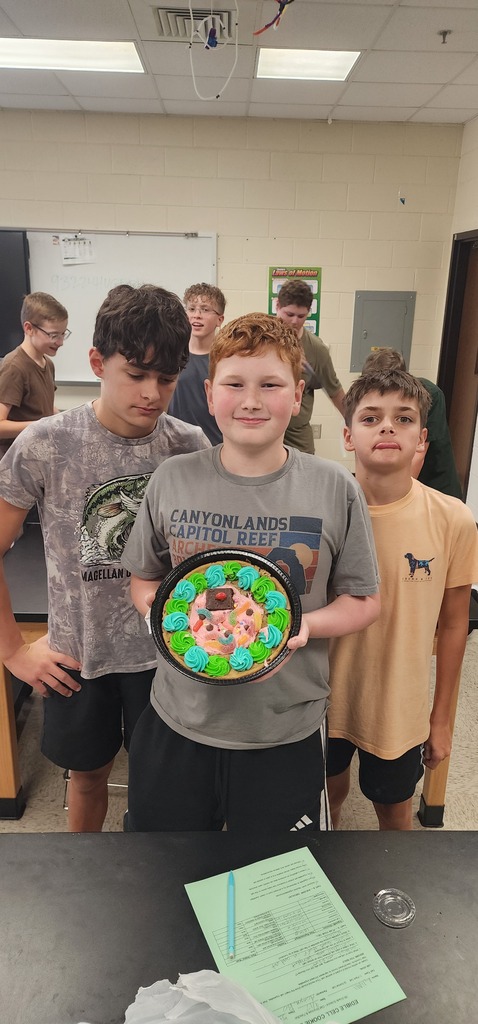 Several boys in a classroom stand around a table; one boy holds a circular art piece.