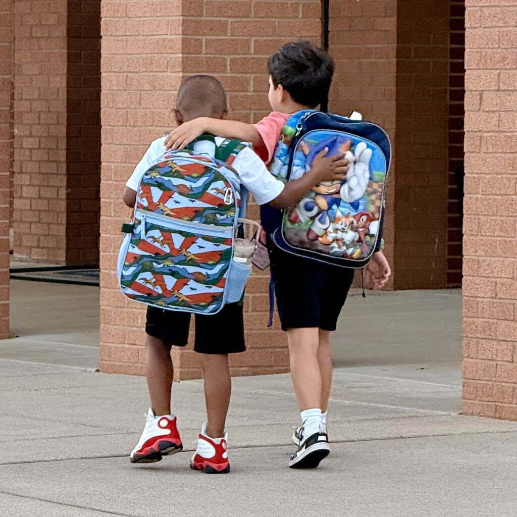 Two kids walking, holding onto each other's shoulders, one with a colorful backpack, the other with a cartoon design.