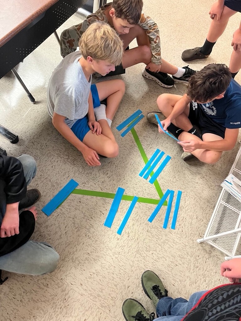 Children sitting on floor, looking at blue and green tape on floor, in a classroom.