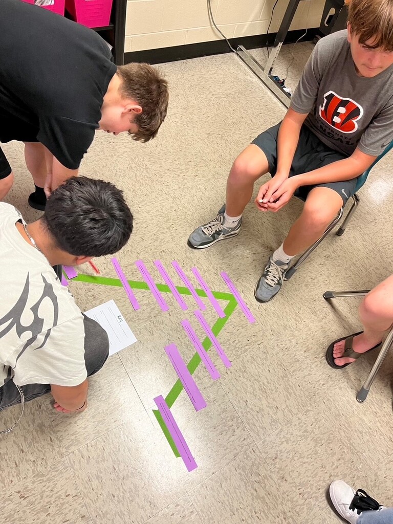 Three boys are on the floor, working on a math activity with purple, green, and yellow paper strips.