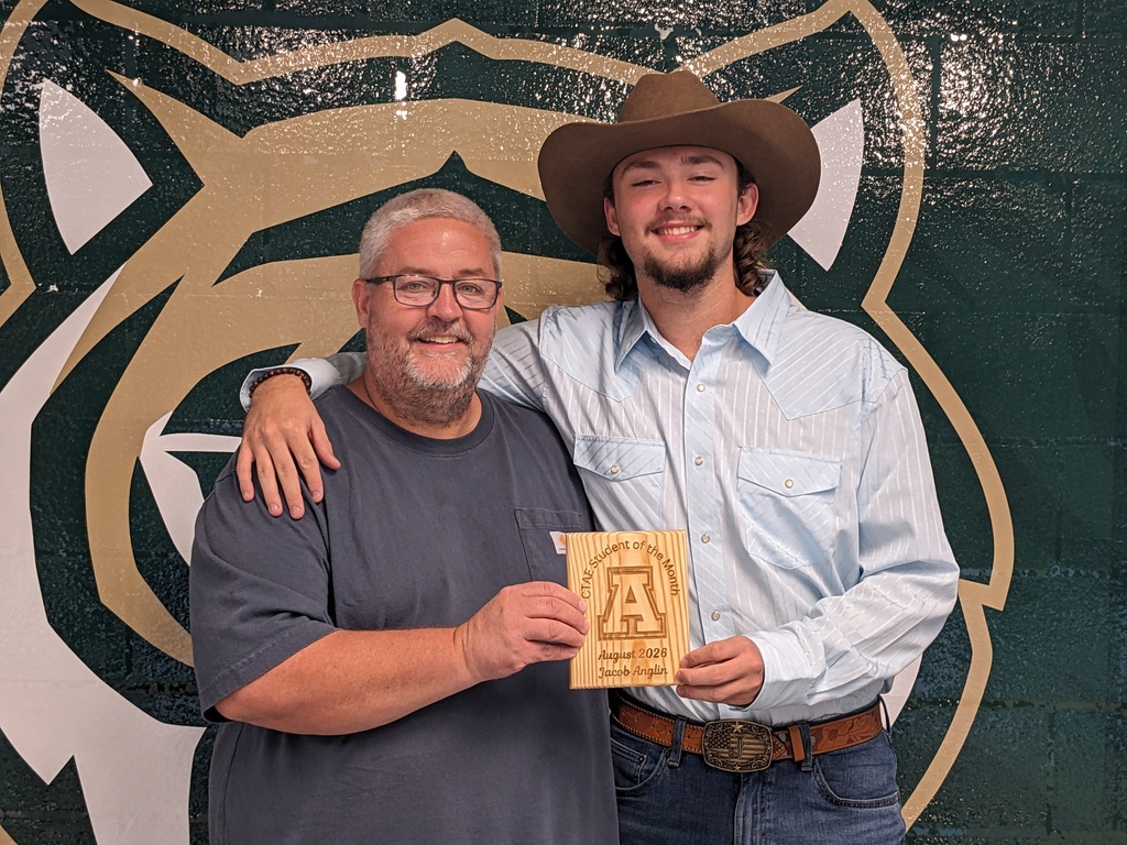 Two men pose for a photo, one in a cowboy hat holding a certificate, with a wall featuring a large animal emblem.