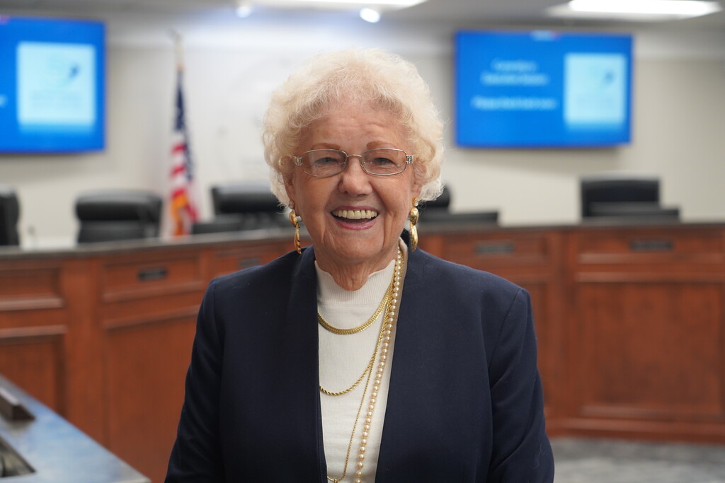 A woman in a dark blue blazer and multiple necklaces stands in an office with a wooden desk.