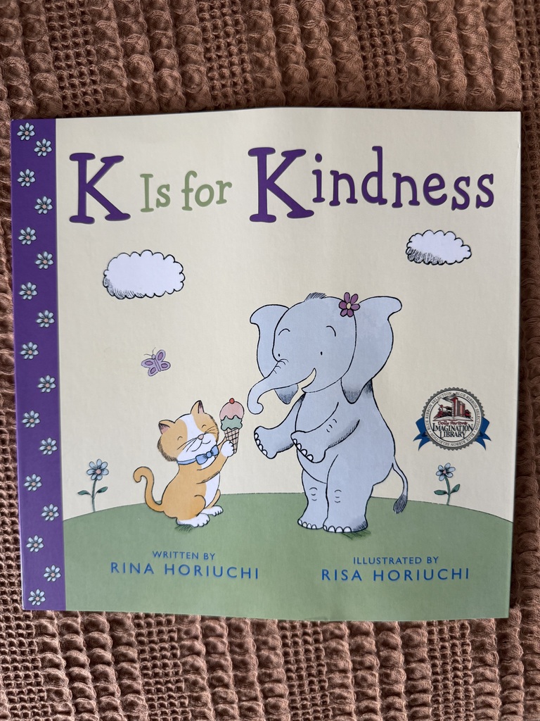A book titled "K Is for Kindness" featuring an elephant and a cat. The elephant holds ice cream, and the cat looks at it. Background is green with flowers and clouds.