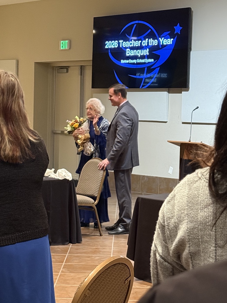 A person presents flowers to a standing woman in a blue dress. A man in a suit stands behind her. An award screen reads "Teacher of the Year Banquet."