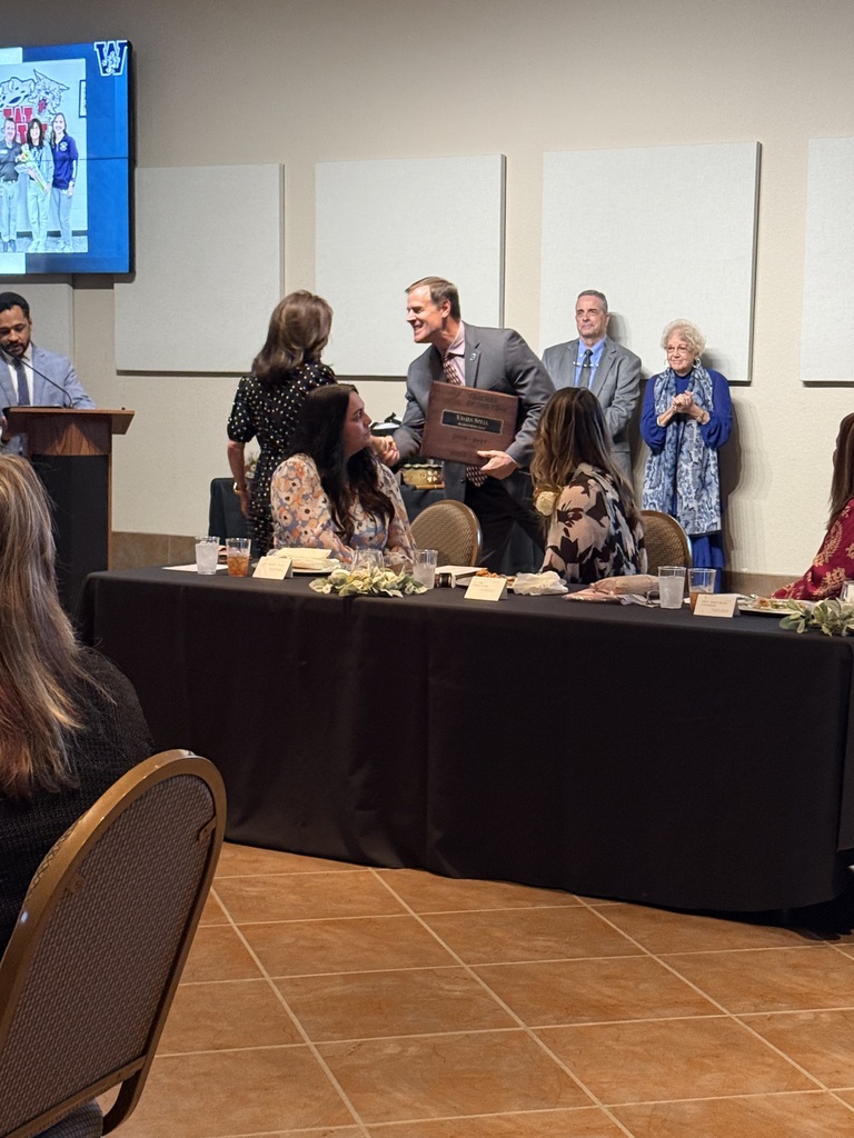 Group in room. Man presenting award to woman. Table with flowers. Blank wall in background.
