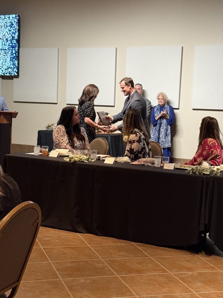 Group of people seated and standing around a long table. One person hands over an award to another.