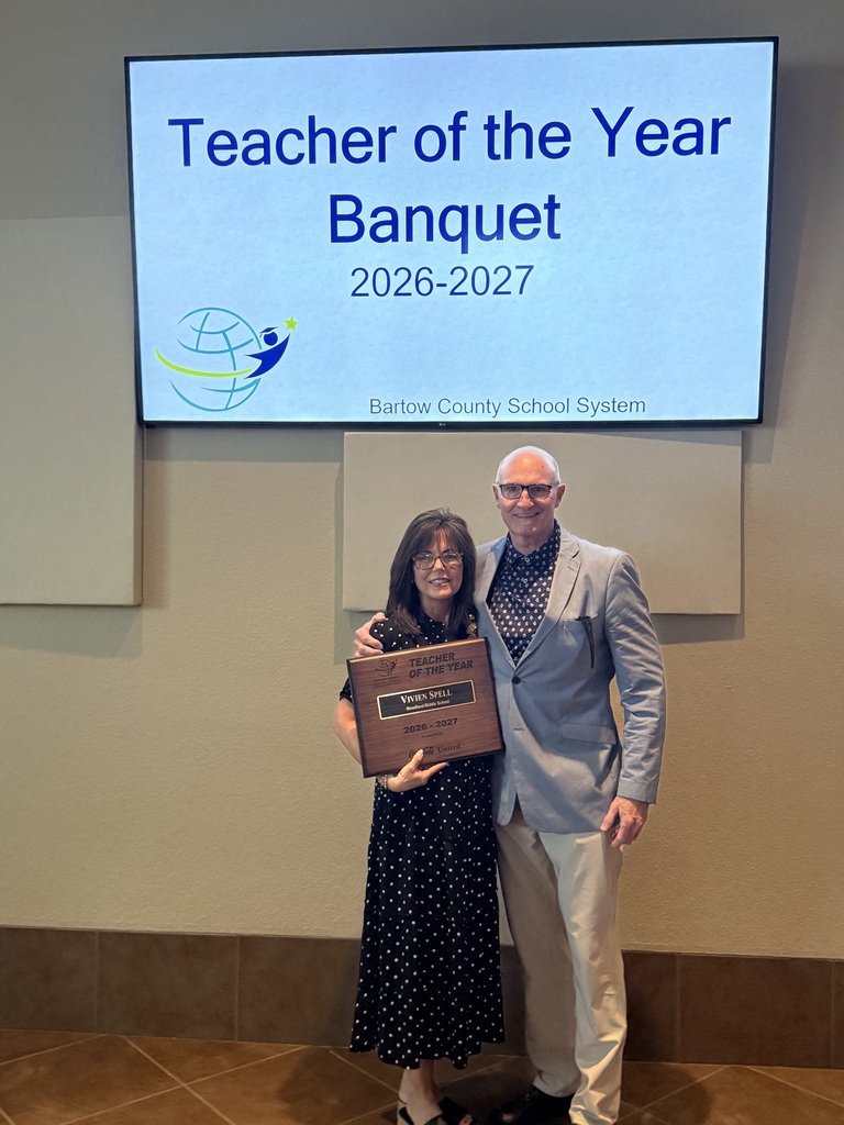 A woman holds an award while standing next to a man in front of a projector screen that reads "Teacher of the Year Banquet 2026-2027".
