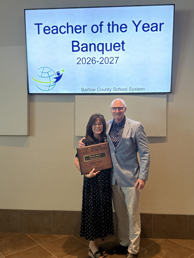 A man and woman pose next to a sign. The woman holds a plaque. The sign reads "Teacher of the Year Banquet 2026-2027."