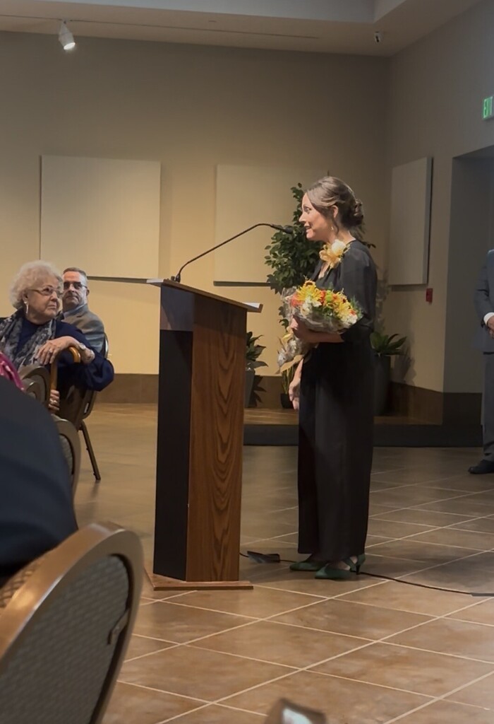 Woman stands at podium with microphone, holding flowers. People seated in chairs in the background.