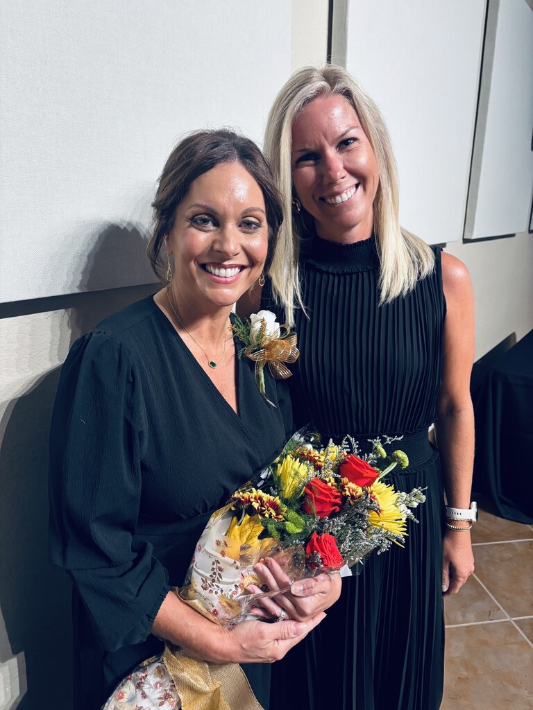 Two women smile, one holding a bouquet of flowers, in a room with a tiled floor.