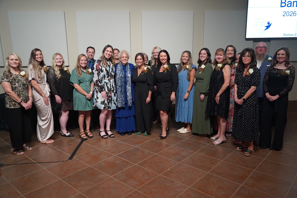 A group of women and men wearing formal attire stand together for a photo, some wearing flowers.