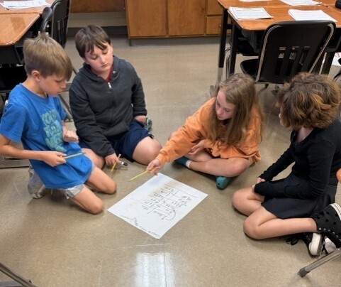 Four children are sitting on the floor in a classroom, with one child holding a drawing.
