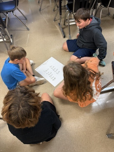 Four children sit on a classroom floor. They are discussing a paper with text and numbers.