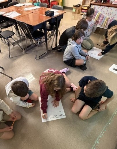 Classroom floor with seven children sitting and standing around, some holding papers and writing.