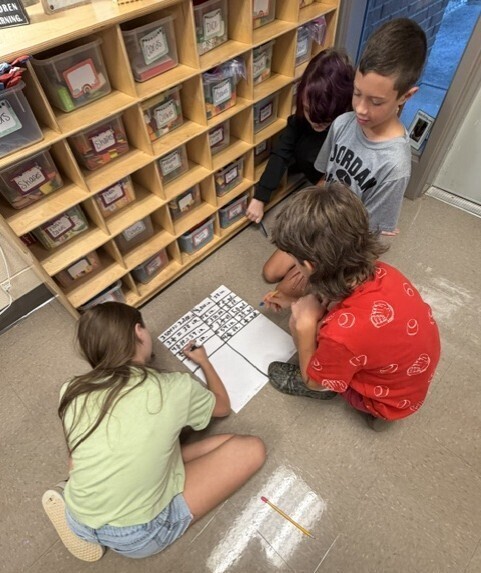 Three children are sitting on the floor, working on a puzzle with a teacher. Behind them is a wooden storage unit with multiple compartments.