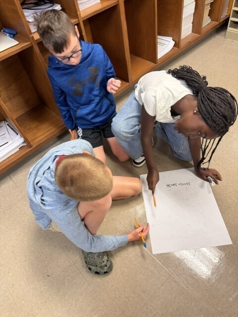 Three children work together on a project, with one writing on a large paper and two others observing.