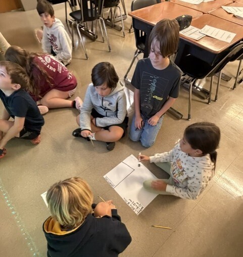 A classroom setting with children sitting on the floor, working on a paper. Two children are standing.