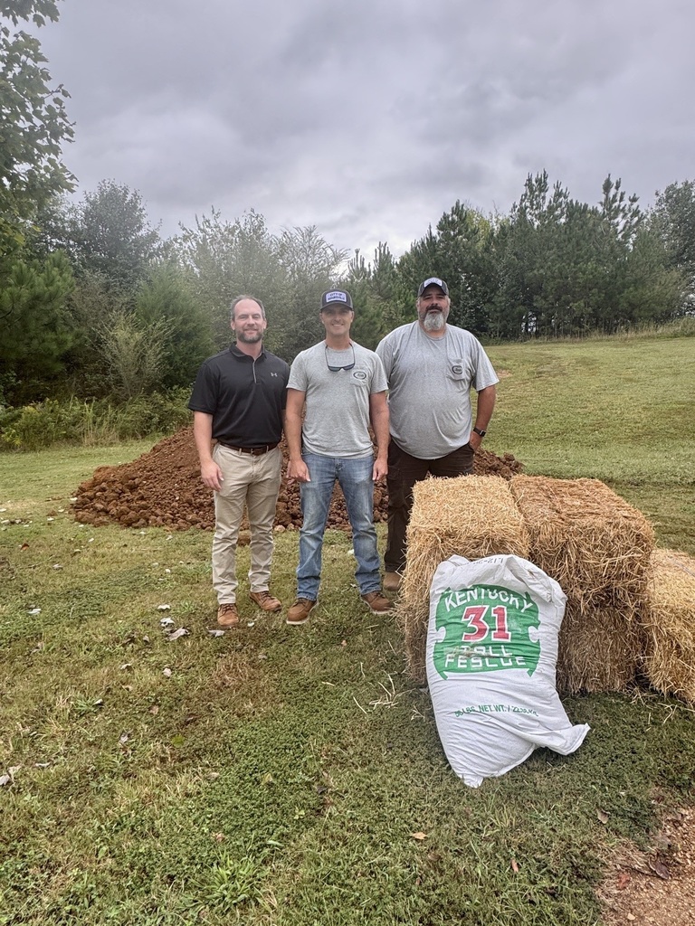 Three men in a field with hay bales, one holding a bag labeled "Kentucky 31 Fall Fescue". Background features trees and a cloudy sky.