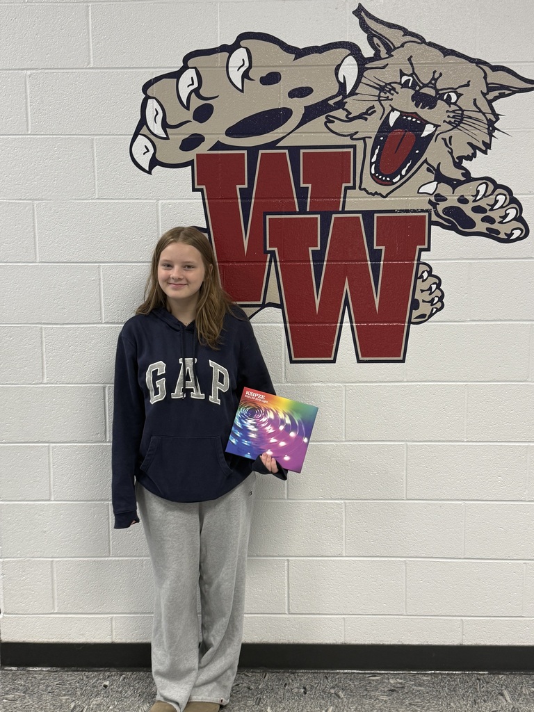 A girl with a blue sweatshirt and gray pants stands before a white wall with a wolf logo.