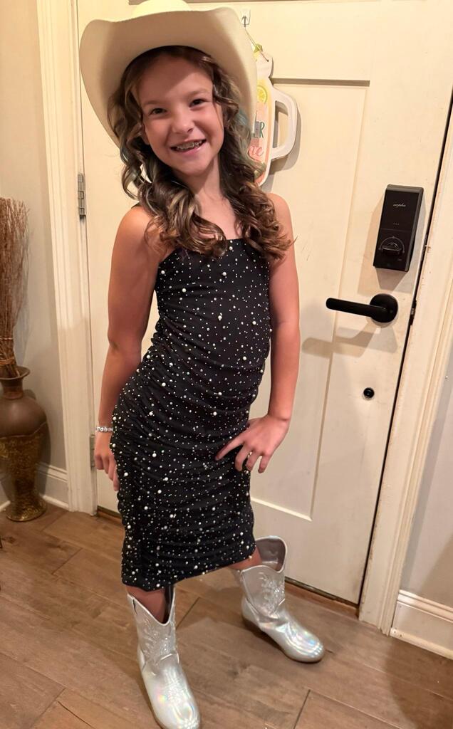 A young girl in a sequined black dress, cowboy hat, and white boots stands near a door. Behind her, a vase and a wall with a lock are visible.