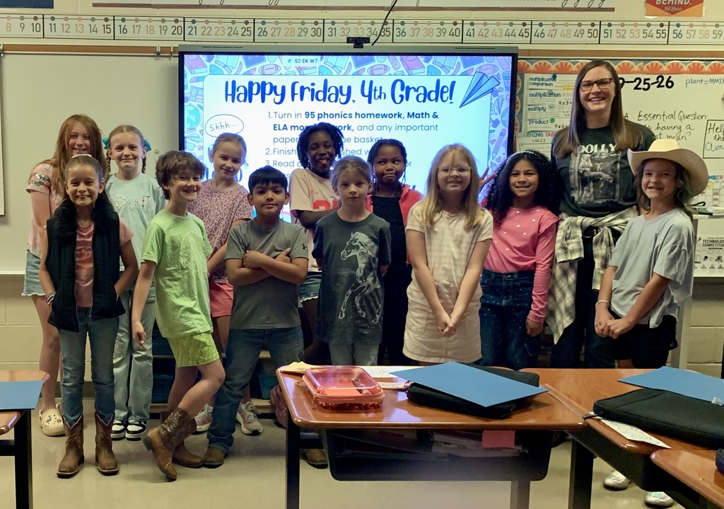 A classroom with children standing around a teacher with a projector screen behind them.