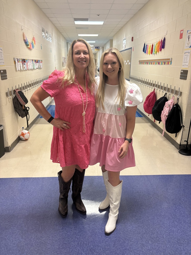 Two women stand in a hallway. One wears a pink dress and boots, the other a pink-and-white dress with boots.
