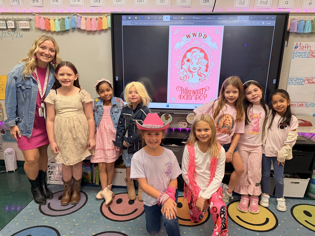 Nine children in matching pink outfits, one wearing a cowboy hat, posing in front of a classroom screen.