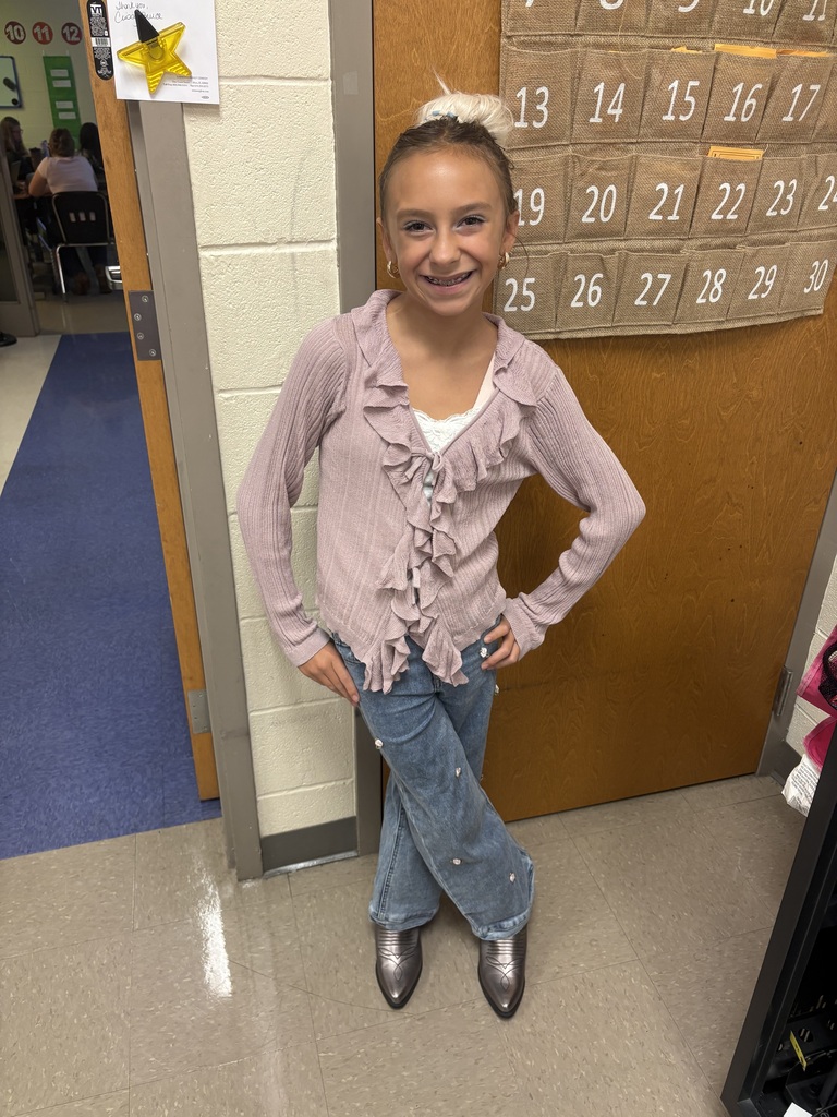 A young girl in pink top and blue jeans stands with hands on hips in a hallway. A calendar is on a brown door behind her.