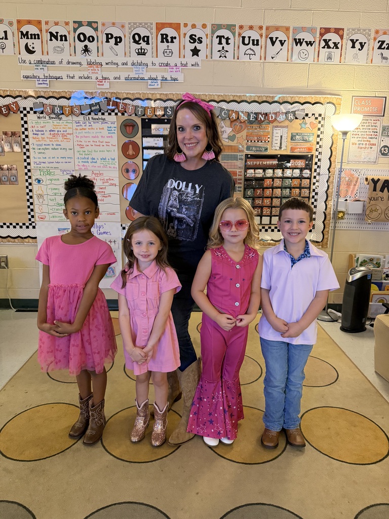 A woman with a pink bow stands with four children in a classroom. The kids wear pink outfits.