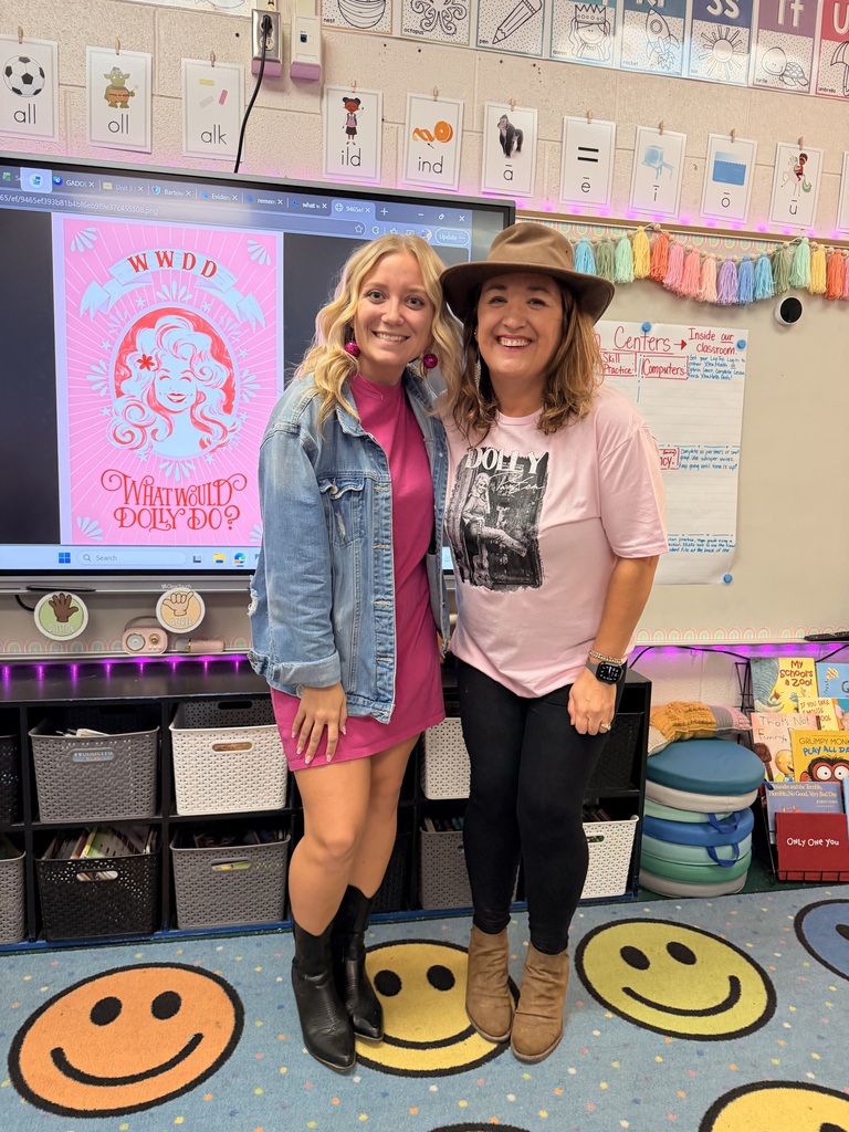 Two women stand in a classroom, smiling for a photo. One wears a pink shirt and a hat; the other wears a denim jacket. Behind them is a large monitor.