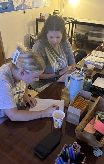 Two women seated at a table, one with a pen, looking at papers and a cup.