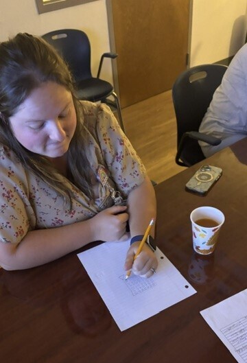 Woman seated at a desk, writing on a piece of paper with a pencil. A cup of tea and a remote control are nearby.