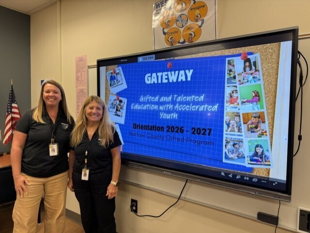 Two women stand in front of a large screen displaying "GATEWAY" and "Accelerated Education." The screen shows images of students and text. An American flag is behind them.