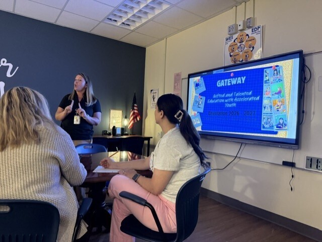 Three people sit in a meeting room. A woman stands in front of a screen displaying a slide.